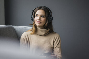 Porträt einer Frau mit Smartphone und schnurlosen Kopfhörern, die sich auf der Couch entspannt und aus dem Fenster schaut - MOEF01837