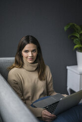 Porträt einer zufriedenen Frau mit hellbraunem Rollkragenpullover, die mit einem Laptop auf der Couch sitzt - MOEF01839