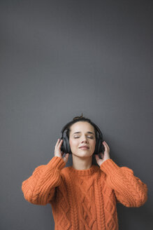 Porträt einer Frau mit orangefarbenem Strickpullover, die an einer grauen Wand lehnt und mit Kopfhörern Musik hört - MOEF01845