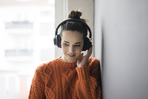 Porträt einer lächelnden Frau mit orangefarbenem Strickpullover, die mit schnurlosen Kopfhörern Musik hört - MOEF01846