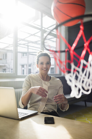 Portrait of smiling freelancer sitting at desk in a loft throwing basketball into hoop stock photo