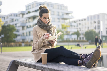 Woman with coffee to go sitting on bench in autumn reading a book - MOEF01862