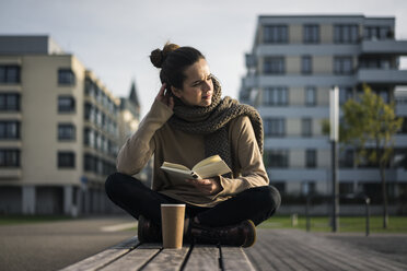 Melancholische Frau mit Kaffee zum Mitnehmen und Buch auf einer Bank im Herbst sitzend - MOEF01863