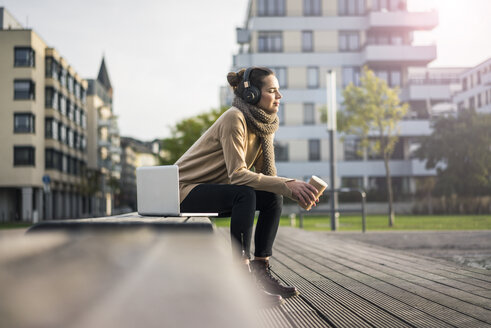Frau mit Kaffee zum Mitnehmen sitzt im Herbst auf einer Bank und hört mit Laptop und schnurlosen Kopfhörern Musik - MOEF01864