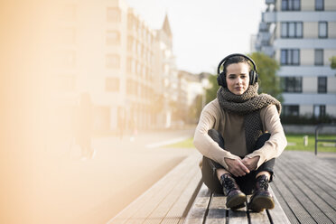 Porträt einer Frau, die im Herbst auf einer Bank sitzt und mit schnurlosen Kopfhörern Musik hört - MOEF01868