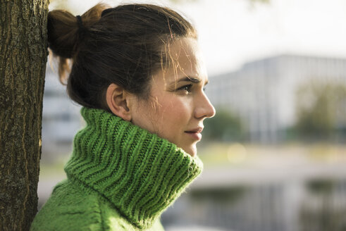 Profil einer entspannten Frau mit grünem Rollkragenpullover, die sich gegen einen Baumstamm lehnt - MOEF01875