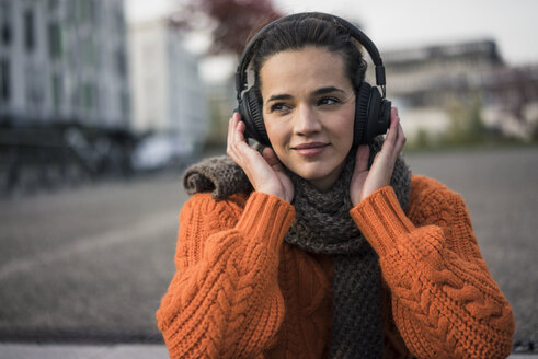 Porträt einer Frau mit Schal und orangefarbenem Strickpullover im Freien, die mit schnurlosen Kopfhörern Musik hört - MOEF01878