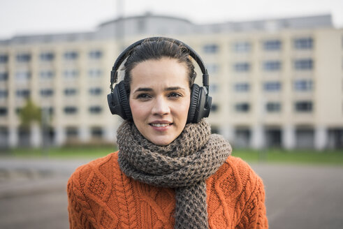 Porträt einer lächelnden Frau mit Schal und orangefarbenem Strickpullover, die mit Kopfhörern Musik hört - MOEF01881