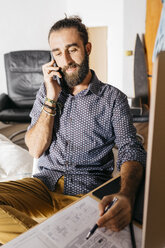 Young architect working at home with blueprints while talking on the phone - JRFF02174