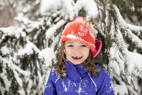 Porträt eines fröhlichen Mädchens in warmer Kleidung, das vor einem verschneiten Baum steht, lizenzfreies Stockfoto