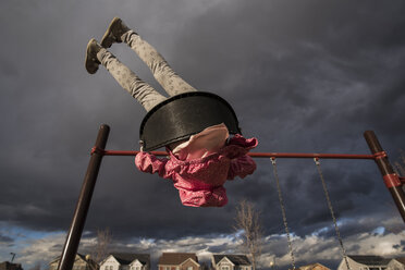 Low angle view of playful girl swinging on swing against stormy clouds at playground - CAVF58020