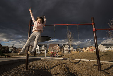 Verspieltes Mädchen springt von der Schaukel gegen stürmische Wolken auf dem Spielplatz - CAVF58022