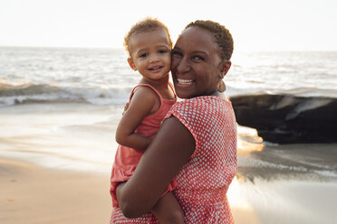 Portrait of mother carrying daughter while standing at beach during sunset - CAVF58035