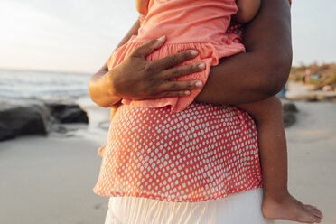 Mittelteil einer schwangeren Mutter, die ihre Tochter trägt, während sie bei Sonnenuntergang am Strand steht - CAVF58036