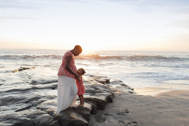 Daughter kissing pregnant mother stomach while standing at beach against sky - CAVF58037