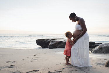 Tochter küsst schwangere Mutter Bauch beim Stehen am Strand bei Sonnenuntergang - CAVF58039