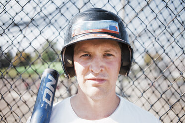 Close-up portrait of confident baseball player wearing sports helmet against chainlink fence at playing field during sunny day - CAVF58085