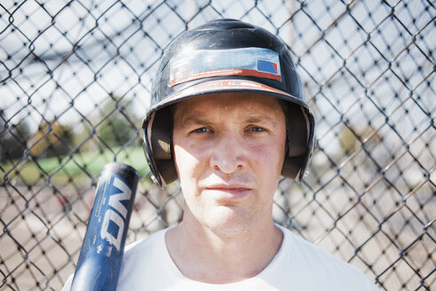 Nahaufnahme eines selbstbewussten Baseballspielers mit Sporthelm vor einem Maschendrahtzaun auf einem Spielfeld an einem sonnigen Tag - CAVF58085