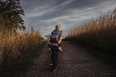 Rückansicht eines Jungen, der seinen Bruder auf einer Landstraße gegen stürmische Wolken huckepack nimmt, lizenzfreies Stockfoto