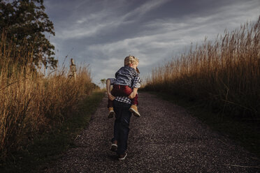 Rückansicht eines Jungen, der seinen Bruder auf einer Landstraße gegen stürmische Wolken huckepack nimmt - CAVF58097