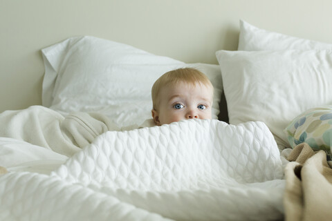 Niedlicher kleiner Junge versteckt sich unter der Bettdecke auf dem Bett zu Hause, lizenzfreies Stockfoto
