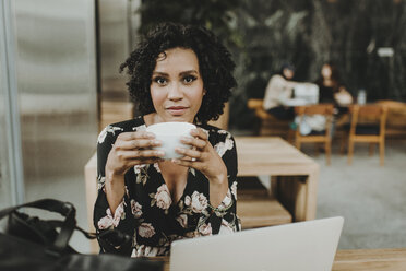 Portrait of woman having coffee while sitting by laptop computer at table in cafe - CAVF58110