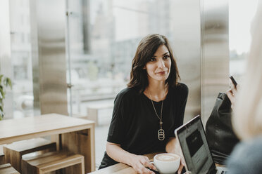 Woman with coffee cup looking away while sitting by laptop computer in cafe - CAVF58113