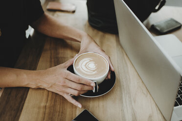 Mittelteil einer Frau, die einen Cappuccino neben einem Laptop auf einem Tisch im Café hält - CAVF58116