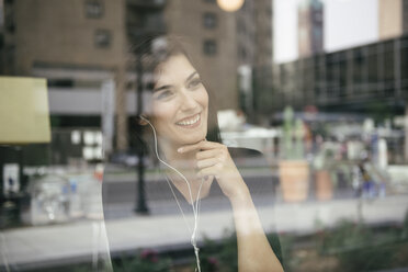 Happy woman listening music while looking through window in cafe seen through glass - CAVF58124