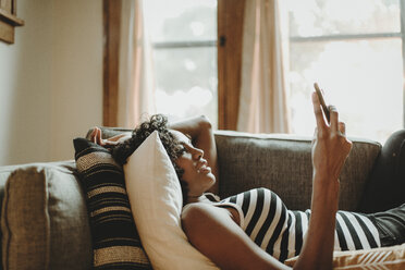 Seitenansicht einer Frau, die ein Smartphone benutzt, während sie zu Hause auf dem Sofa liegt - CAVF58154