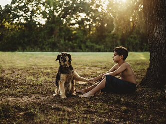 Side view of shirtless teenage boy sitting with dog on field - CAVF58173