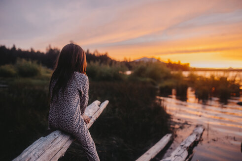 Verspieltes Mädchen, das bei Sonnenuntergang auf Treibholz sitzt und die Aussicht betrachtet - CAVF58176