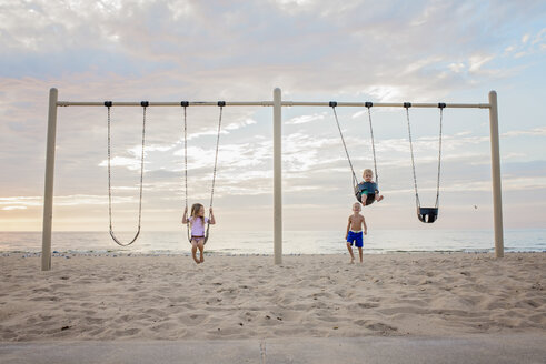 Geschwister spielen auf Schaukeln am Strand gegen bewölkten Himmel bei Sonnenuntergang - CAVF58262
