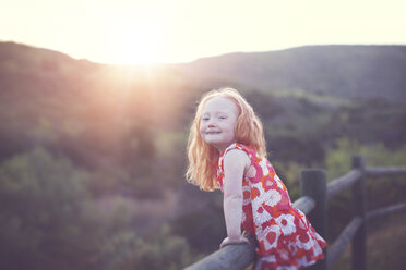 Porträt eines verspielten Mädchens, das sich bei Sonnenuntergang an ein Geländer mit Bergen lehnt - CAVF58277
