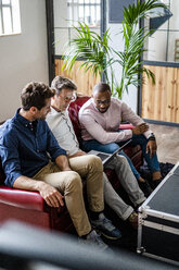 Three businessmen using laptop sitting on sofa in loft office - GIOF04970