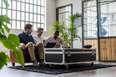 Three businessmen using laptop sitting on sofa in loft office - GIOF04971