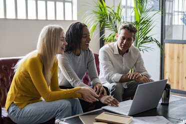 Happy business team using laptop sitting on couch in loft office - GIOF04995