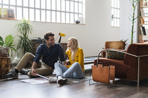 Smiling businessman and businesswoman sitting on the floor discussing documents in loft office - GIOF05005