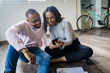 Smiling businessman and businesswoman sitting on the floor with cell phone and documents - GIOF05031