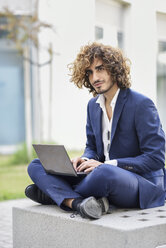 Porträt eines jungen Geschäftsmannes mit lockigem Haar und blauem Anzug, der auf einer Bank im Freien sitzt und einen Laptop benutzt - JSMF00650