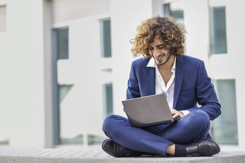 Lächelnder junger Geschäftsmann mit lockigem Haar und blauem Anzug sitzt auf einer Bank im Freien und benutzt einen Laptop, lizenzfreies Stockfoto