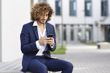 Smiling young businessman with curly hair wearing blue suit sitting on bench outdoors looking at cell phone - JSMF00655