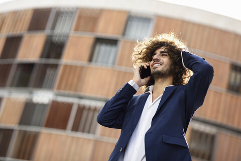 Porträt eines jungen, lächelnden, modischen Geschäftsmannes mit lockigem Haar am Telefon, lizenzfreies Stockfoto