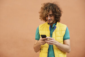 Lächelnder junger Mann mit lockigem Haar und gelber Weste schaut auf sein Handy - JSMF00674