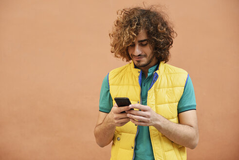 Smiling young man with curly hair wearing yellow waistcoat looking at cell phone - JSMF00674