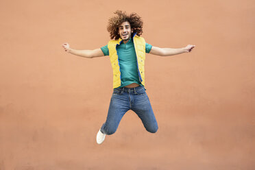 Portrait of smiling young man with curly hair wearing yellow waistcoat jumping in the air - JSMF00676