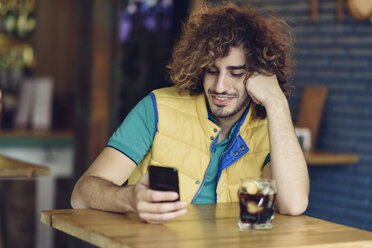 Smiling young man with beard and curly hair looking at cell phone in a pub - JSMF00688