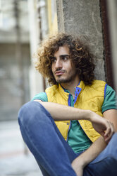 Portrait of young man with beard and curly hair wearing yellow waistcoat watching something - JSMF00690