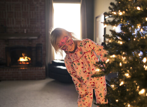 Mädchen mit Gesichtsbemalung, das im Wohnzimmer stehend einen beleuchteten Weihnachtsbaum betrachtet, lizenzfreies Stockfoto