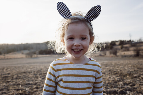 Porträt eines niedlichen, fröhlichen Mädchens, das ein Stirnband mit Kaninchenohren trägt und bei Sonnenuntergang auf einem Feld steht, lizenzfreies Stockfoto